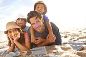 Smiling family of parents and two young boys using Nöz's yellow, blue, orange, and purple reef-safe sunscreen on a sunny day at the beach.